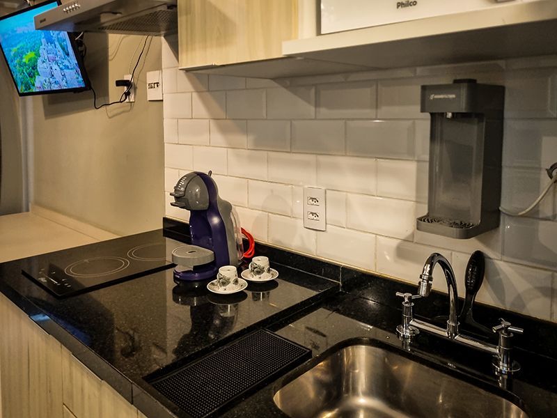 Bancada de cozinha moderna com fogão preto, cafeteira, pia e TV de parede ao fundo.
