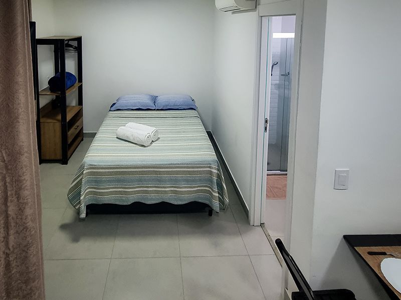 Quarto pequeno com uma cama de solteiro, prateleira e porta para um banheiro.