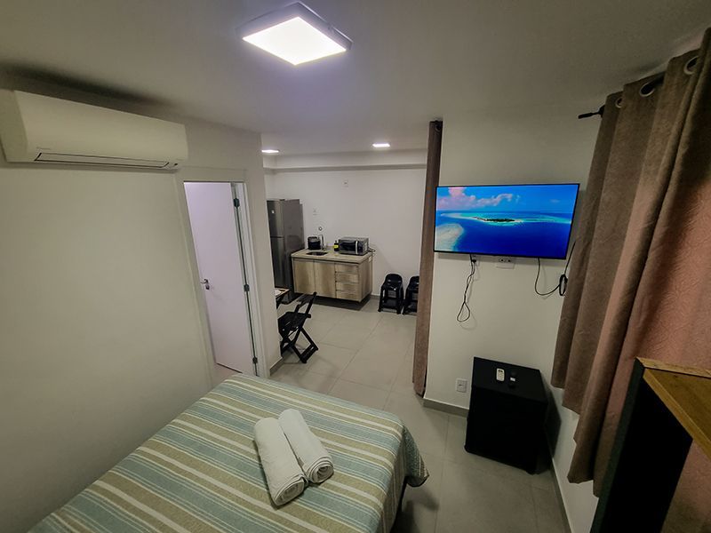 Quarto pequeno com cama, TV de parede, escrivaninha e cortinas ao lado da porta que dá para outro cômodo.