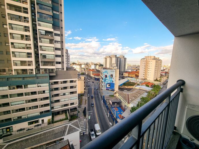 Vista da varanda para uma rua da cidade com prédios altos, trânsito e céu azul ao pôr do sol.