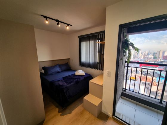 Quarto pequeno com cama, mesa de cabeceira e porta para varanda com vista para a cidade.