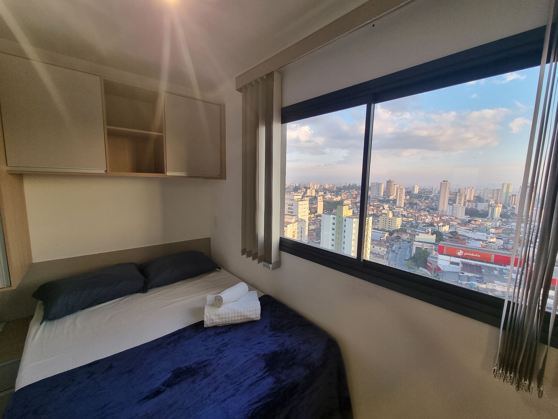 Quarto pequeno com cama e janela ampla com vista para o horizonte da cidade ao entardecer.