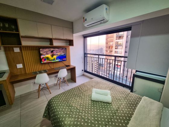 Quarto estúdio moderno com varanda, escrivaninha, TV e vista para a cidade através de portas de vidro deslizantes.