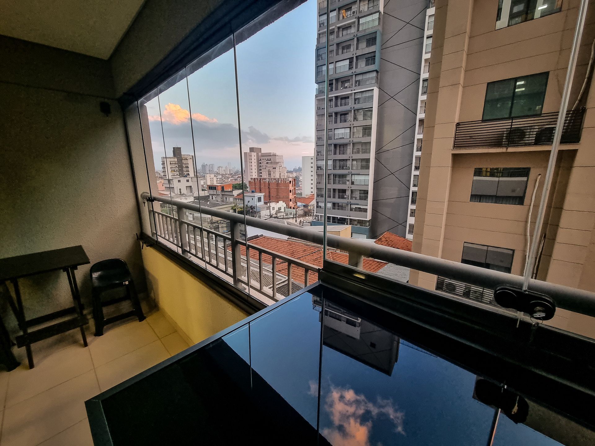 Vista da cidade ao pôr do sol a partir de uma varanda, com uma mesa e cadeira pretas em primeiro plano.