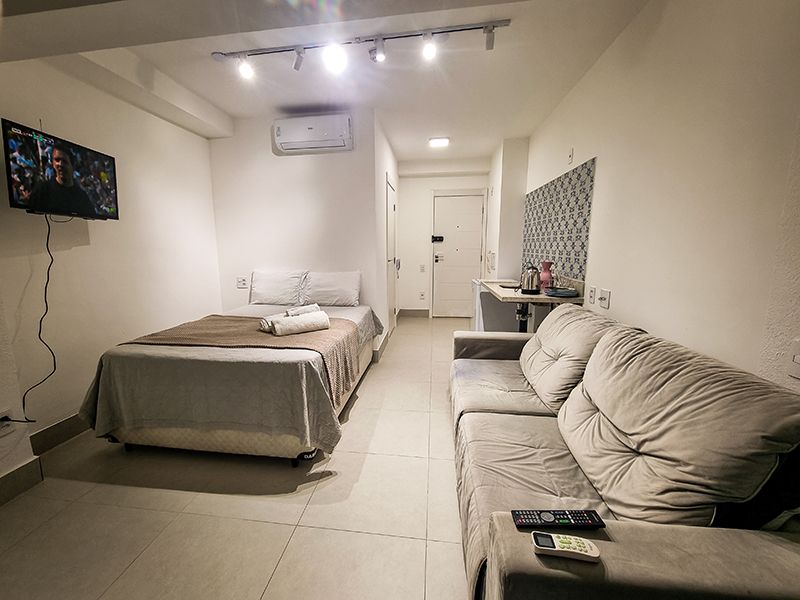 Quarto estúdio luminoso com cama, sofá, TV de parede e escrivaninha em um ambiente minimalista.