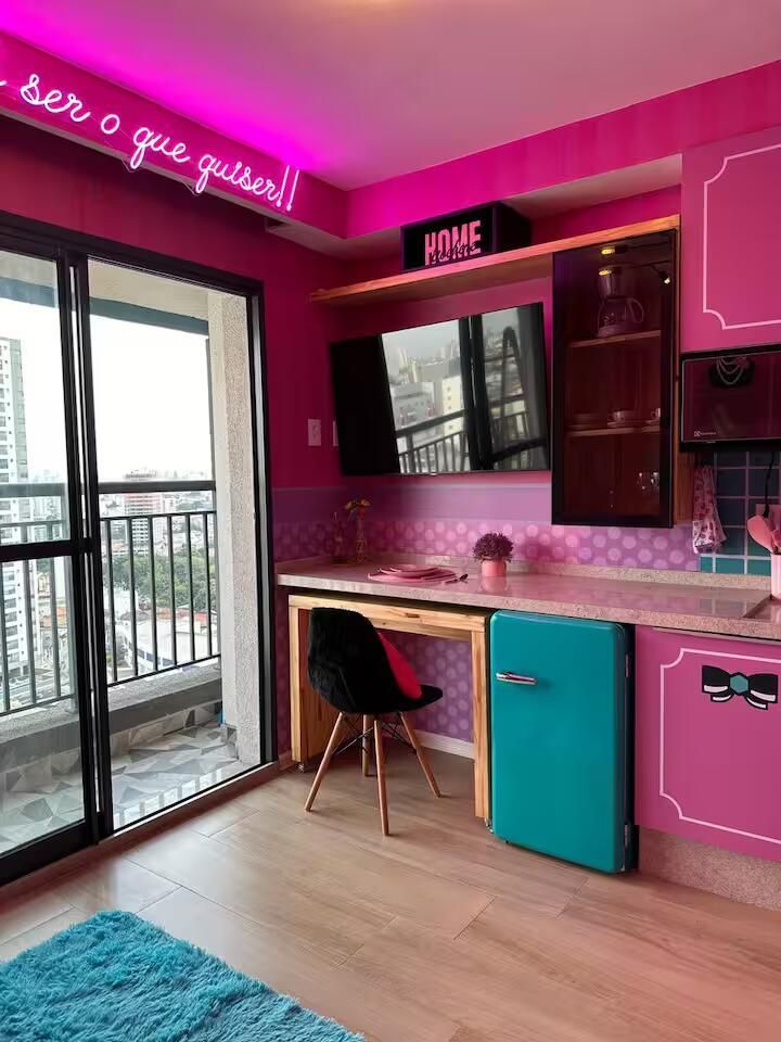 Cantinho de cozinha rosa vibrante com detalhes em azul-petróleo, escrivaninha, armários e portas de vidro deslizantes que dão para uma varanda.
