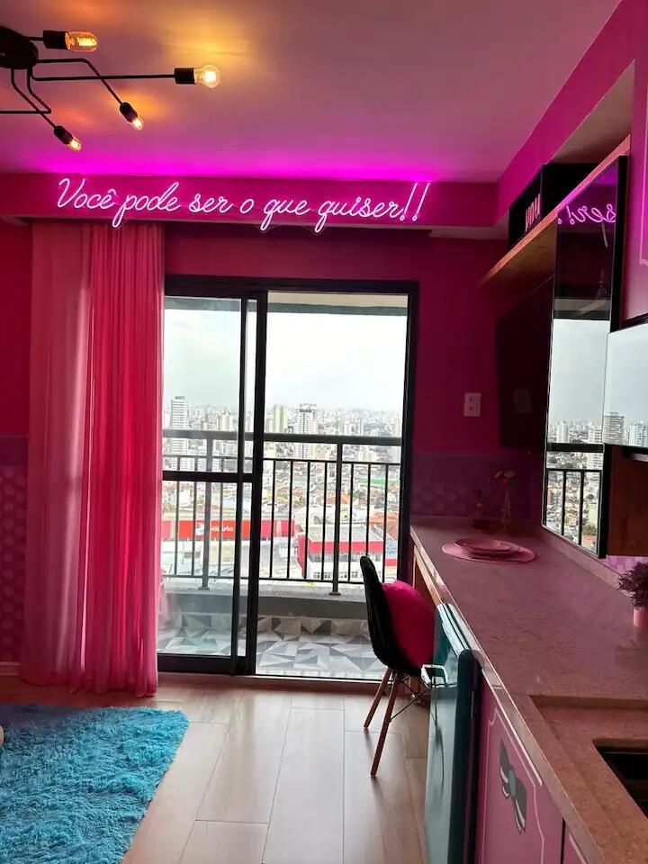 Quarto iluminado em rosa com letreiro de neon na parede, portas de varanda, escrivaninha, cadeira e cortinas.