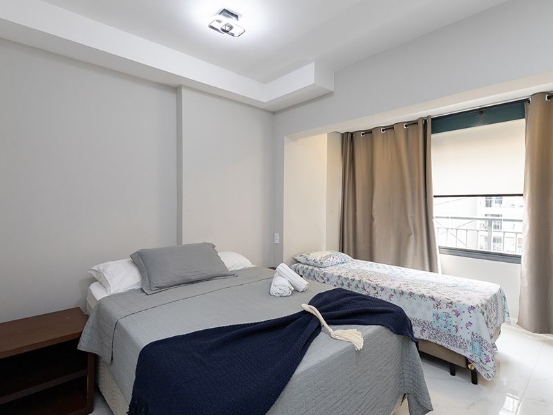 Quarto iluminado com duas camas desarrumadas, um banco escuro e uma janela com cortinas.