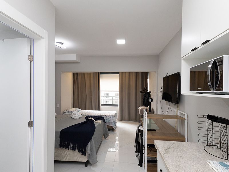 Quarto compacto estilo hotel com duas camas, escrivaninha, TV e cozinha compacta.