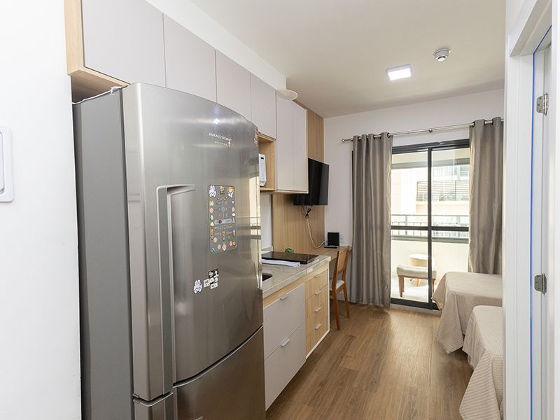 Apartamento estúdio compacto com geladeira prateada, cozinha compacta, cama e portas de varanda.