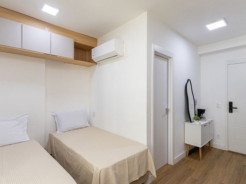 Quarto pequeno e iluminado com duas camas de solteiro, armários brancos, ar-condicionado de parede e uma pequena escrivaninha perto da porta.