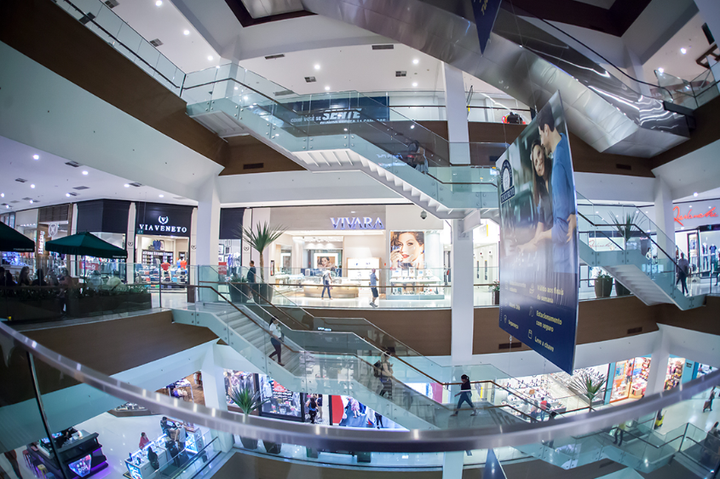 Interior de um shopping center de vários andares com escadas rolantes, guarda-corpos de vidro e vitrines.