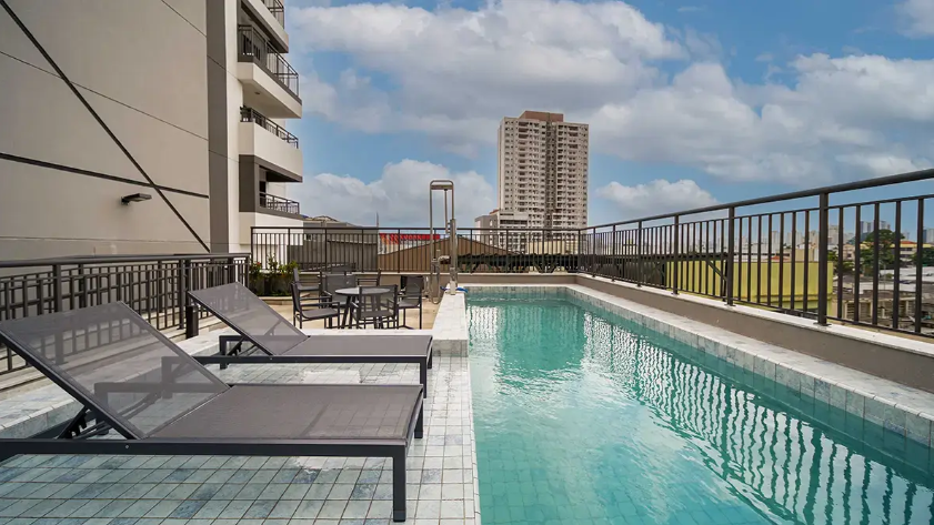 Piscina na cobertura com espreguiçadeiras e vista para os prédios da cidade sob um céu parcialmente nublado.