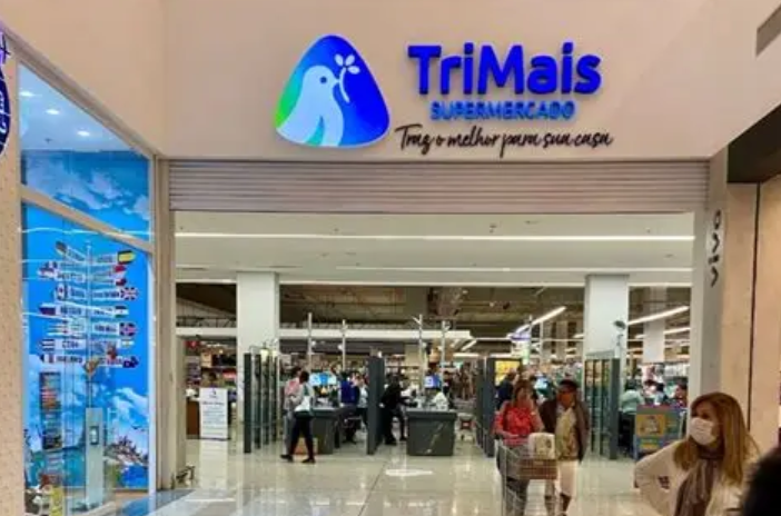 Entrada do supermercado TriMais com clientes e placa azul brilhante.