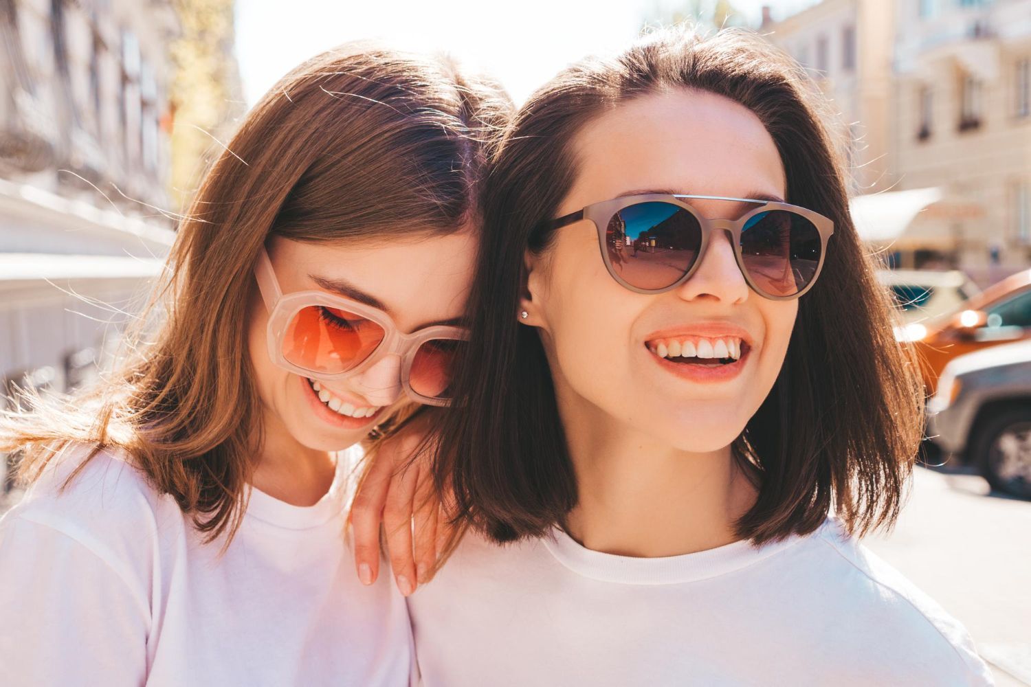 Two women smiling, wearing sunglasses, outside. One has an arm around the other.