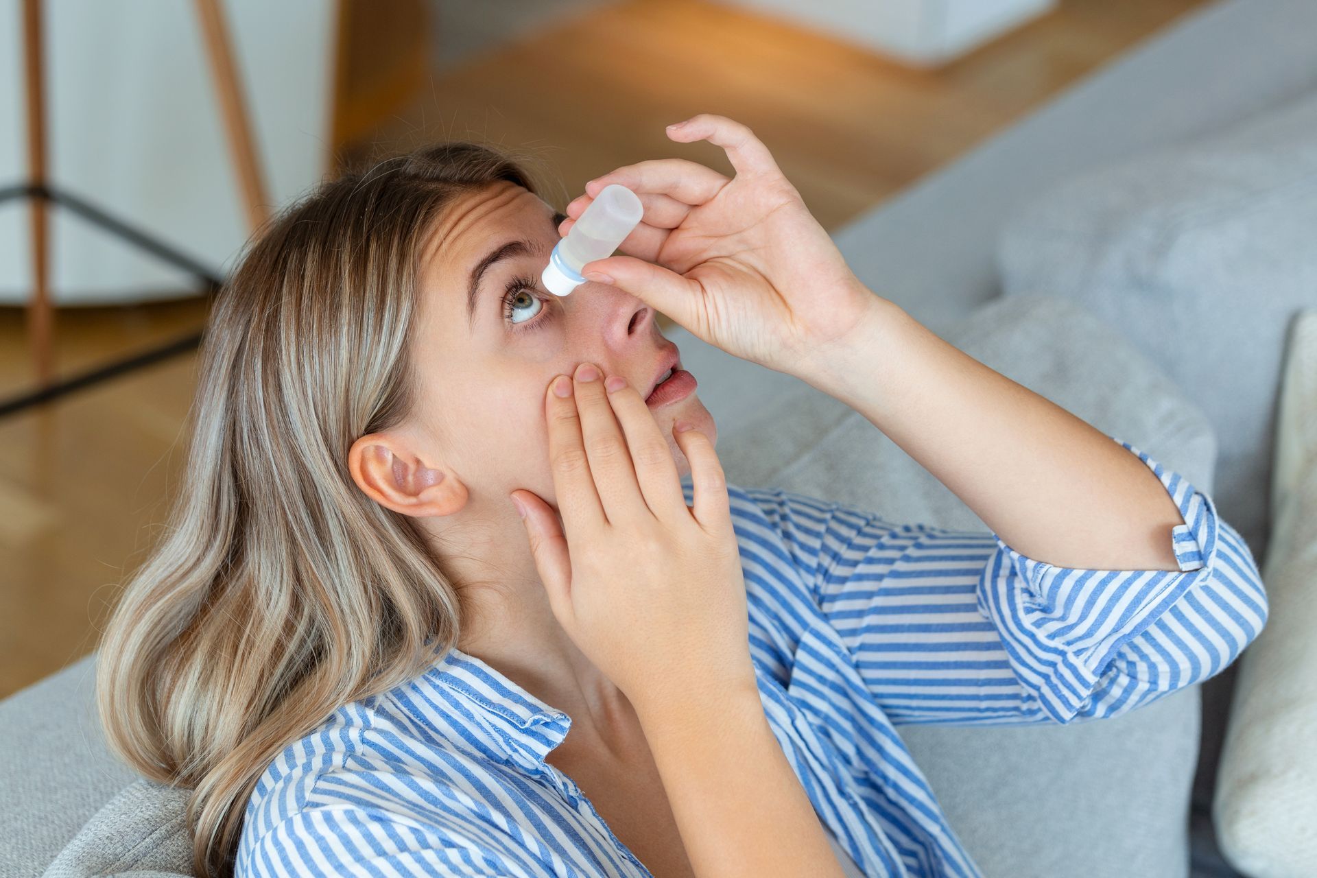Woman applying eye drops while sitting on a couch, wearing a blue striped shirt.