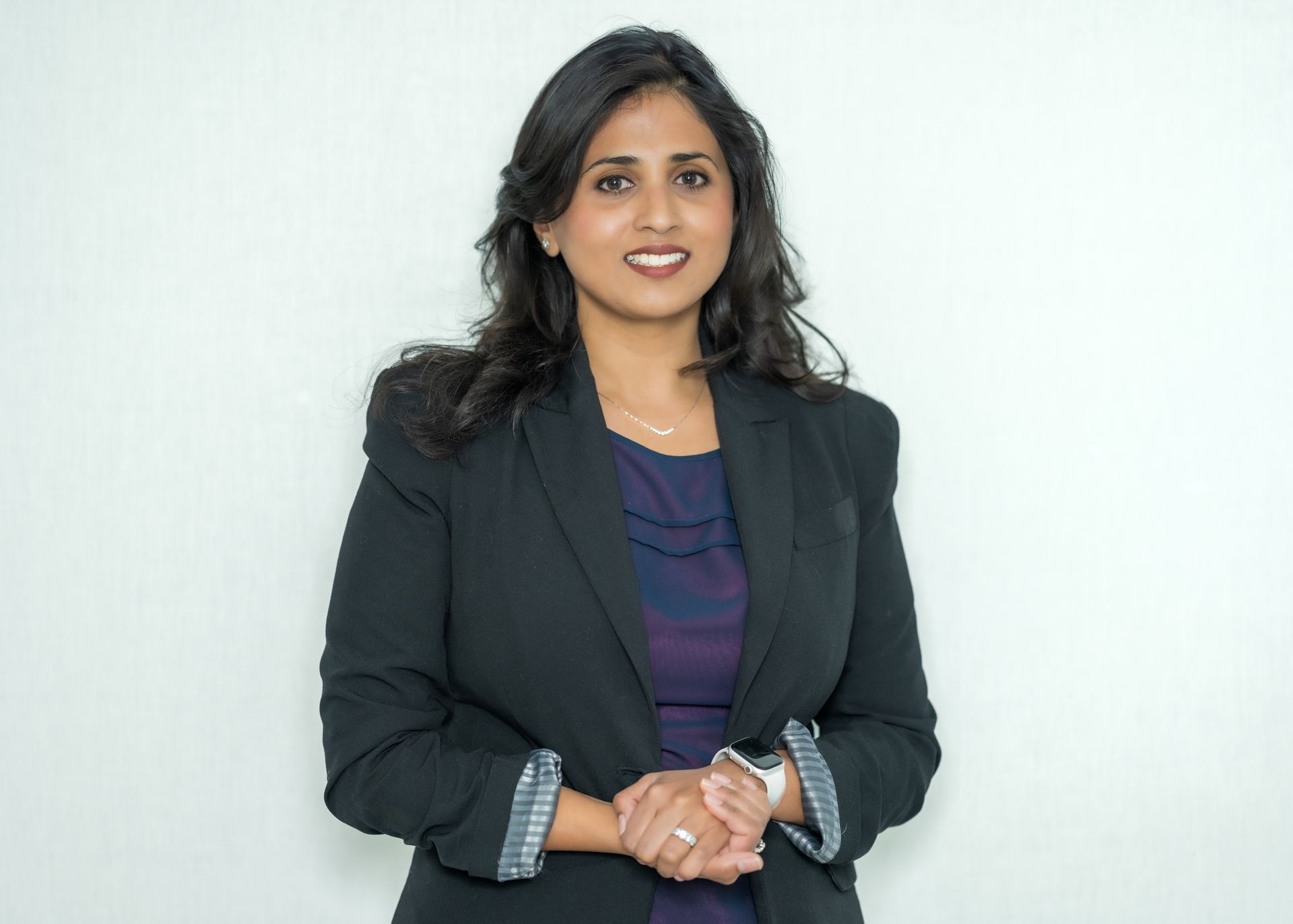 Woman in a dark blazer standing against a plain white background, hands clasped and smiling.
