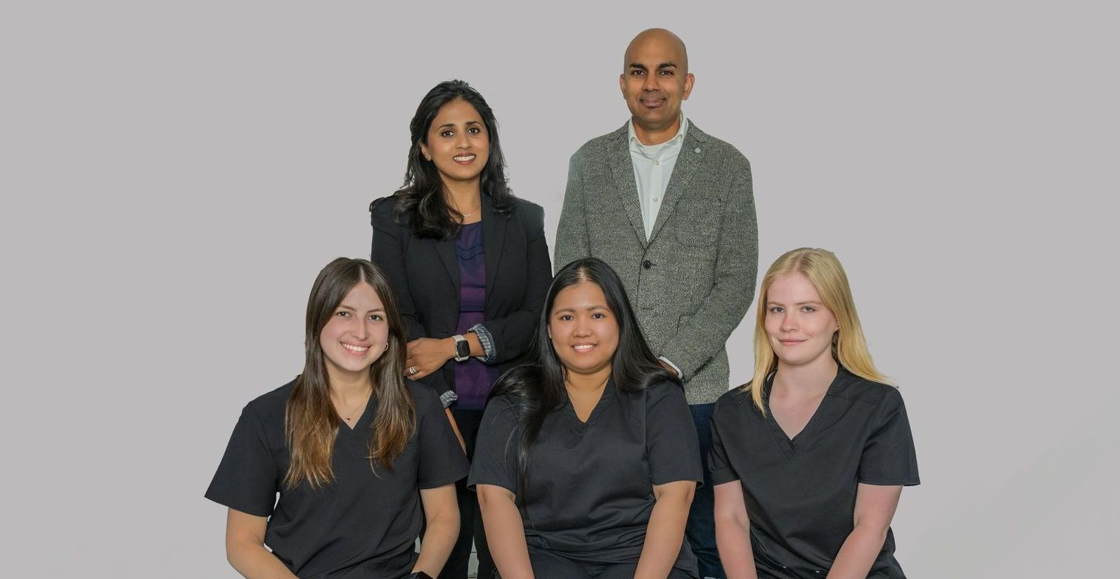 Five coworkers posing together against a plain gray background, two standing behind three seated in front.