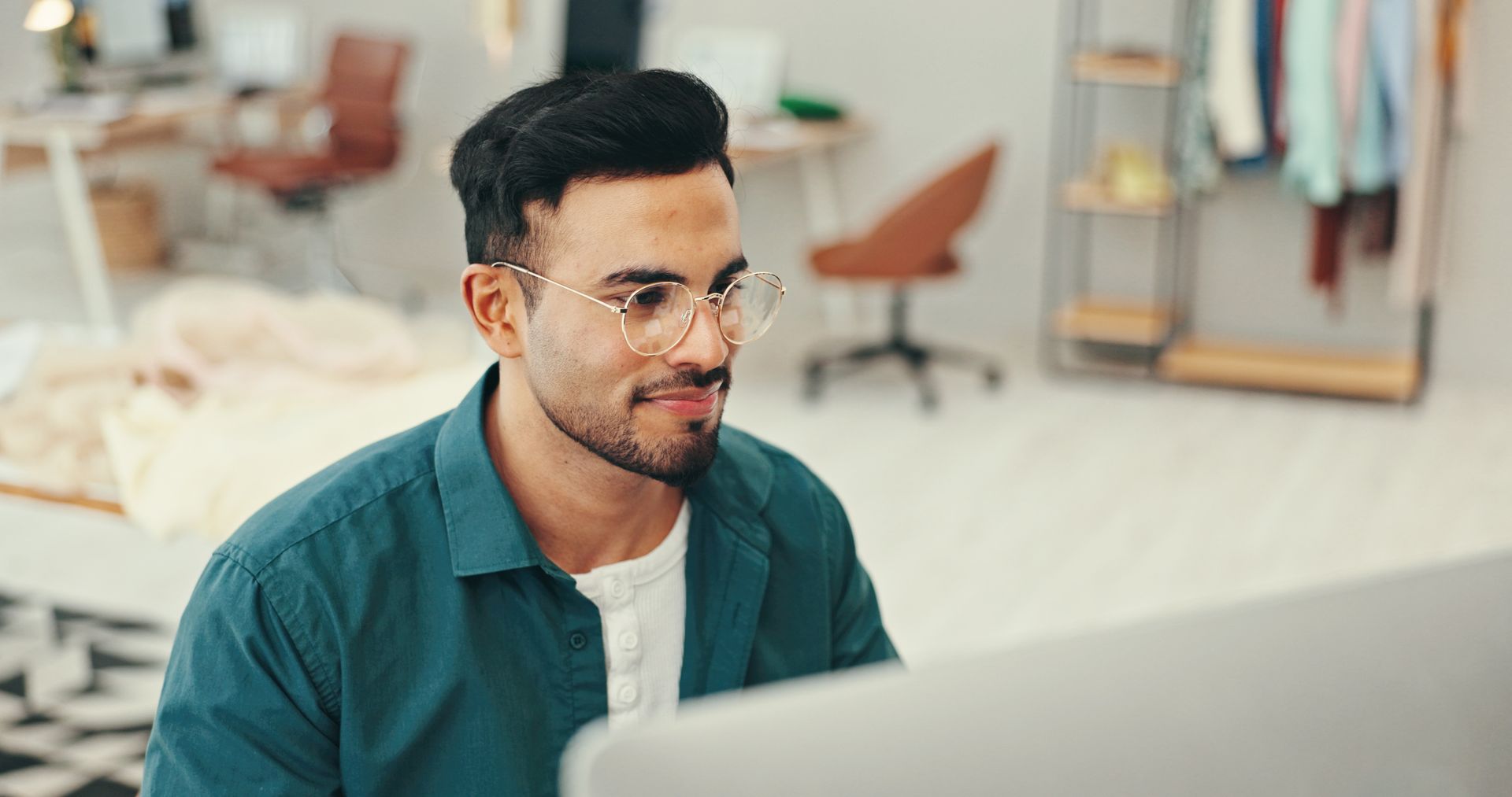 Man smiling at a laptop in a bright office with clothes racks in the background
