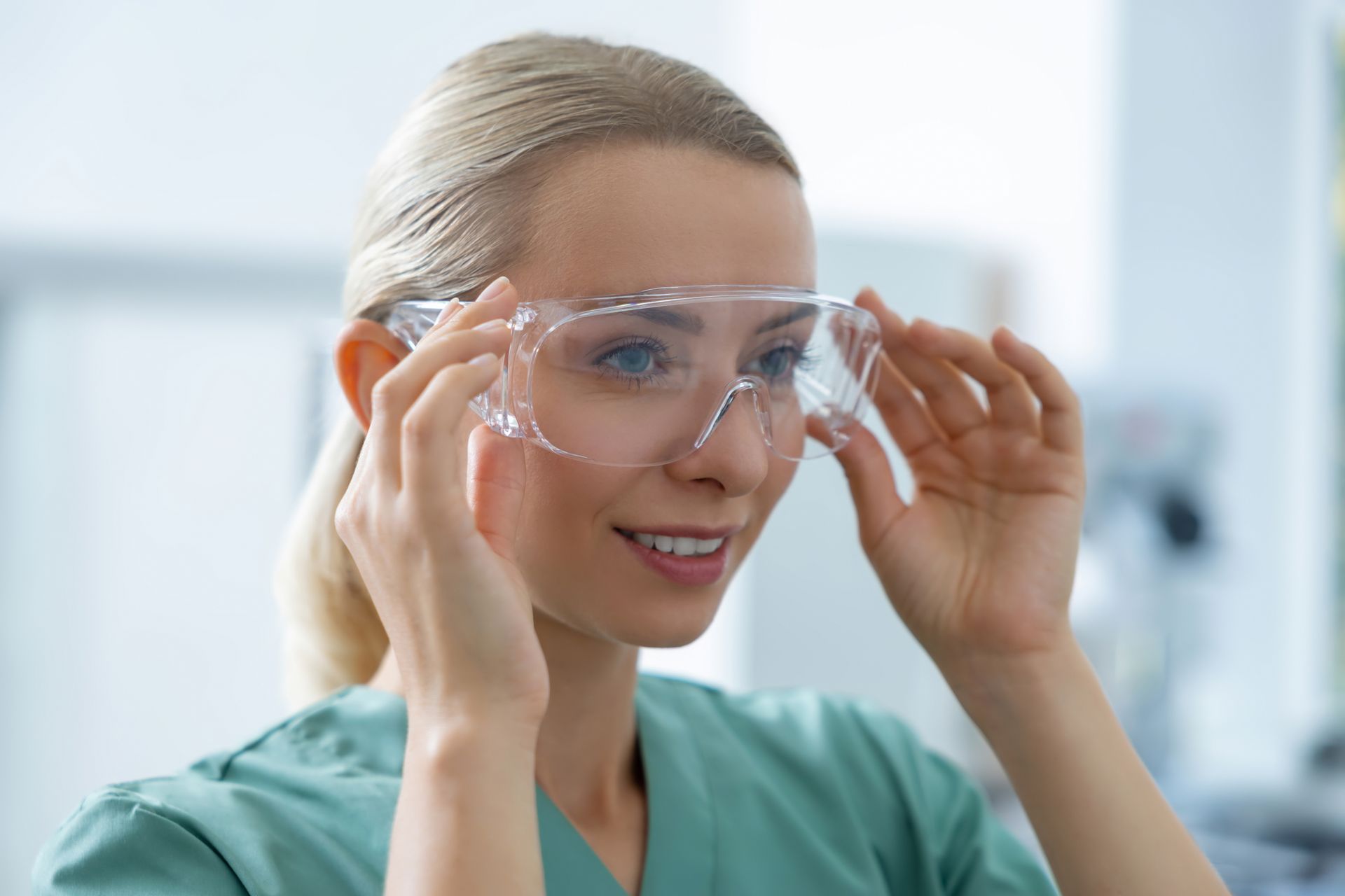Person in green scrubs adjusting clear safety goggles in a bright lab setting
