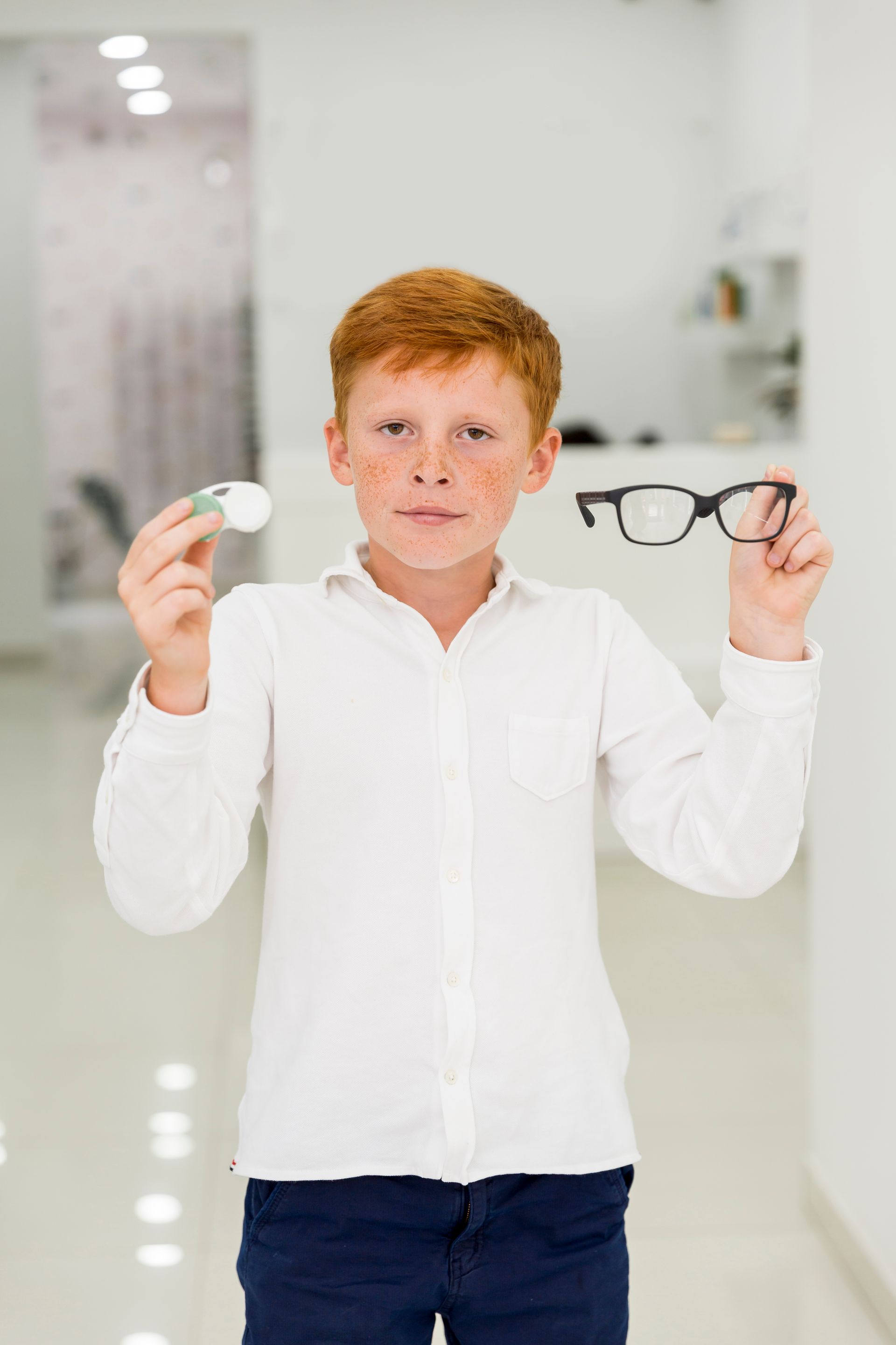 Boy in a white shirt holding a watch in one hand and eyeglasses in the other indoors.