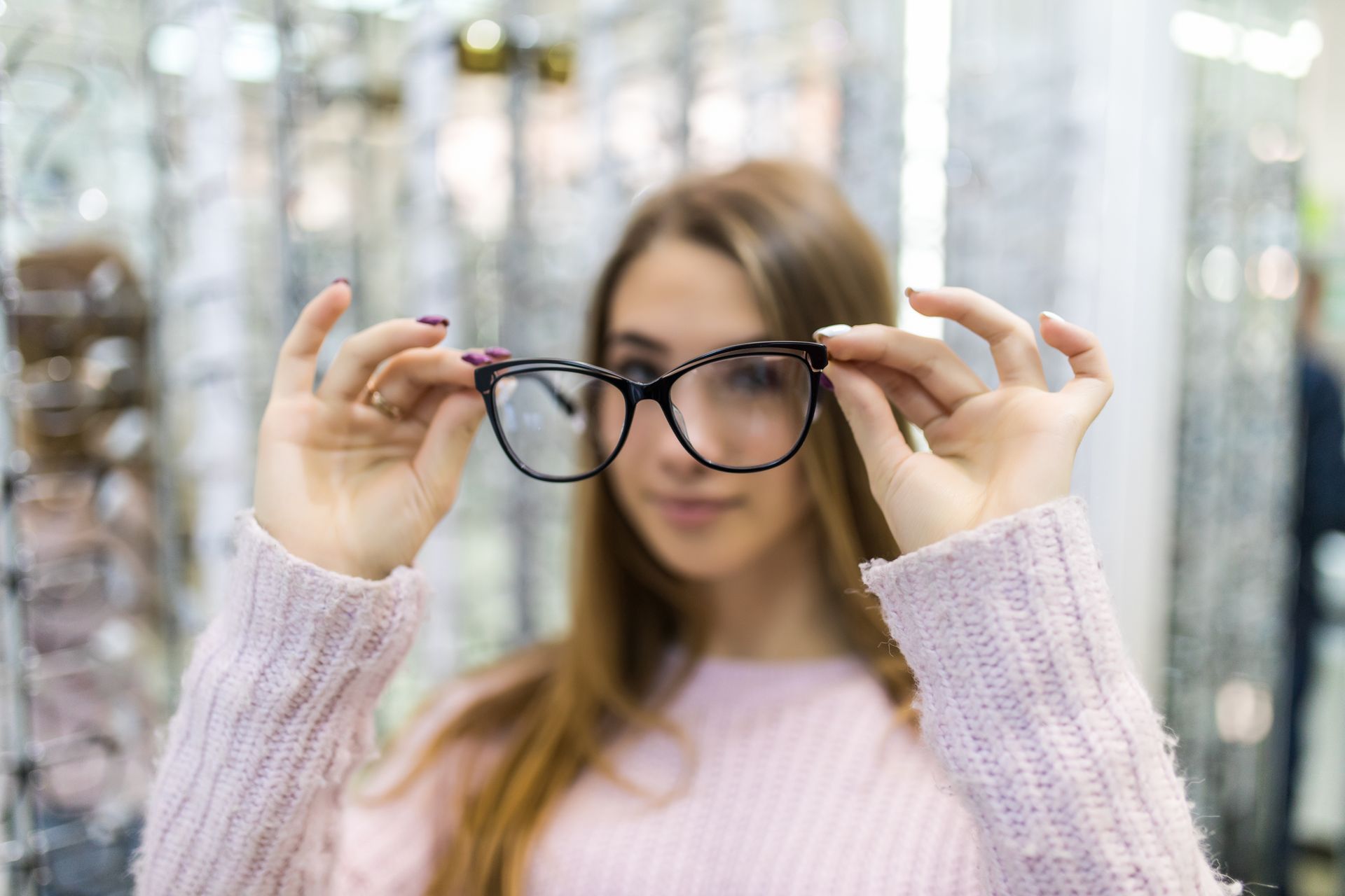 Person holding black eyeglasses in front of face, wearing a light pink sweater in a bright indoor setting