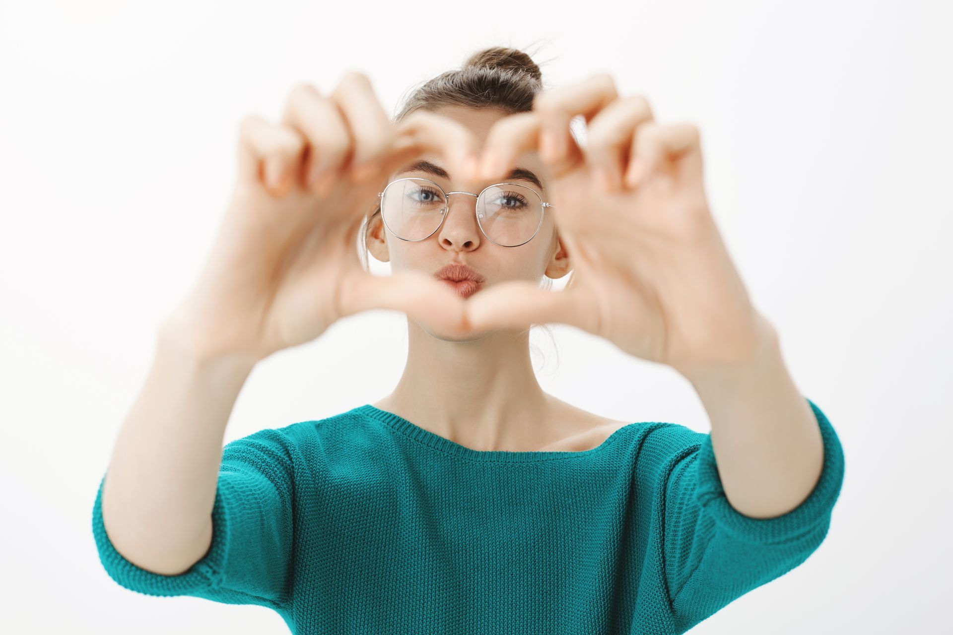 Person holding eyeglasses up to the camera, wearing a teal sweater against a white background