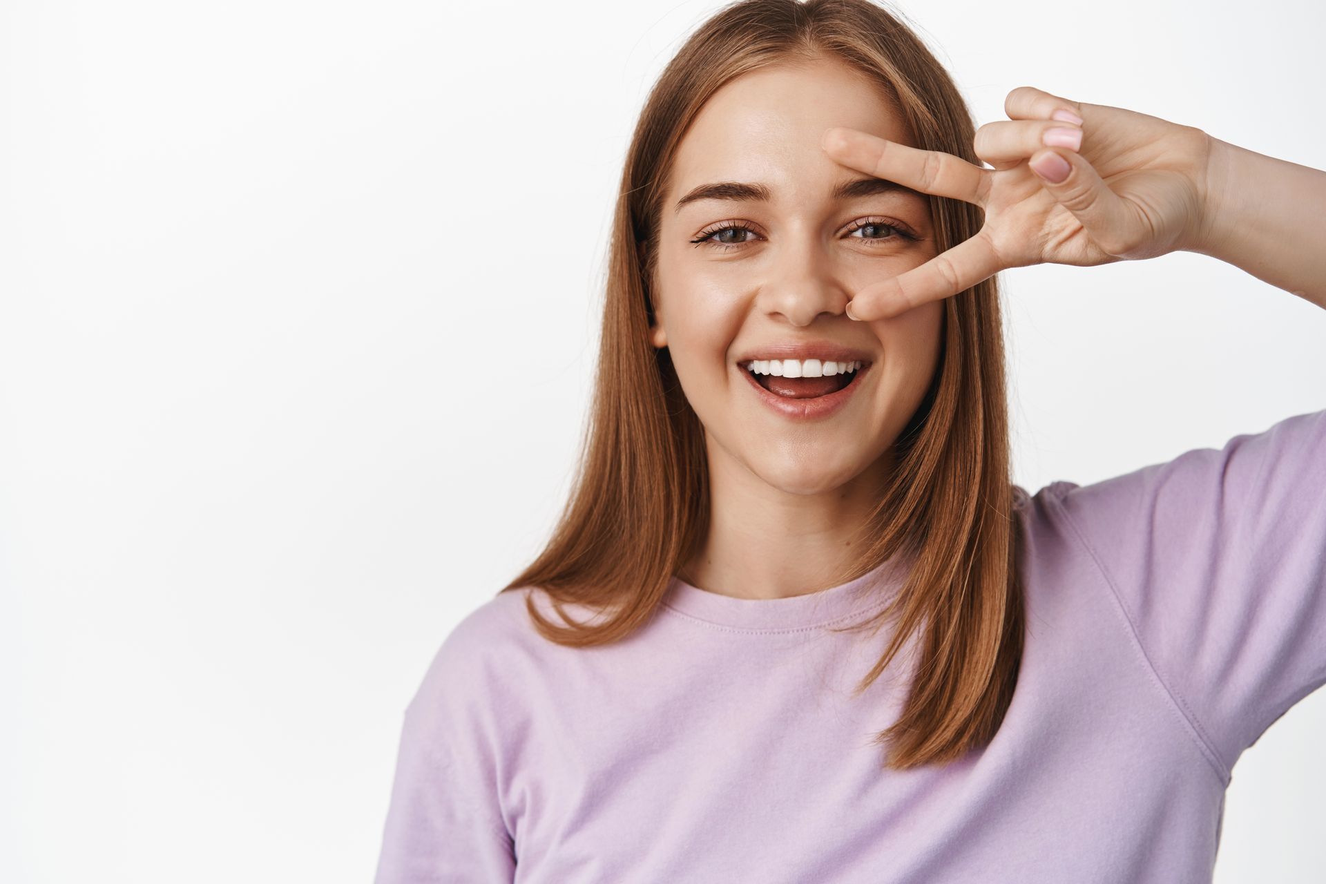 Smiling woman in a lavender shirt making a peace sign near her eye against a white background