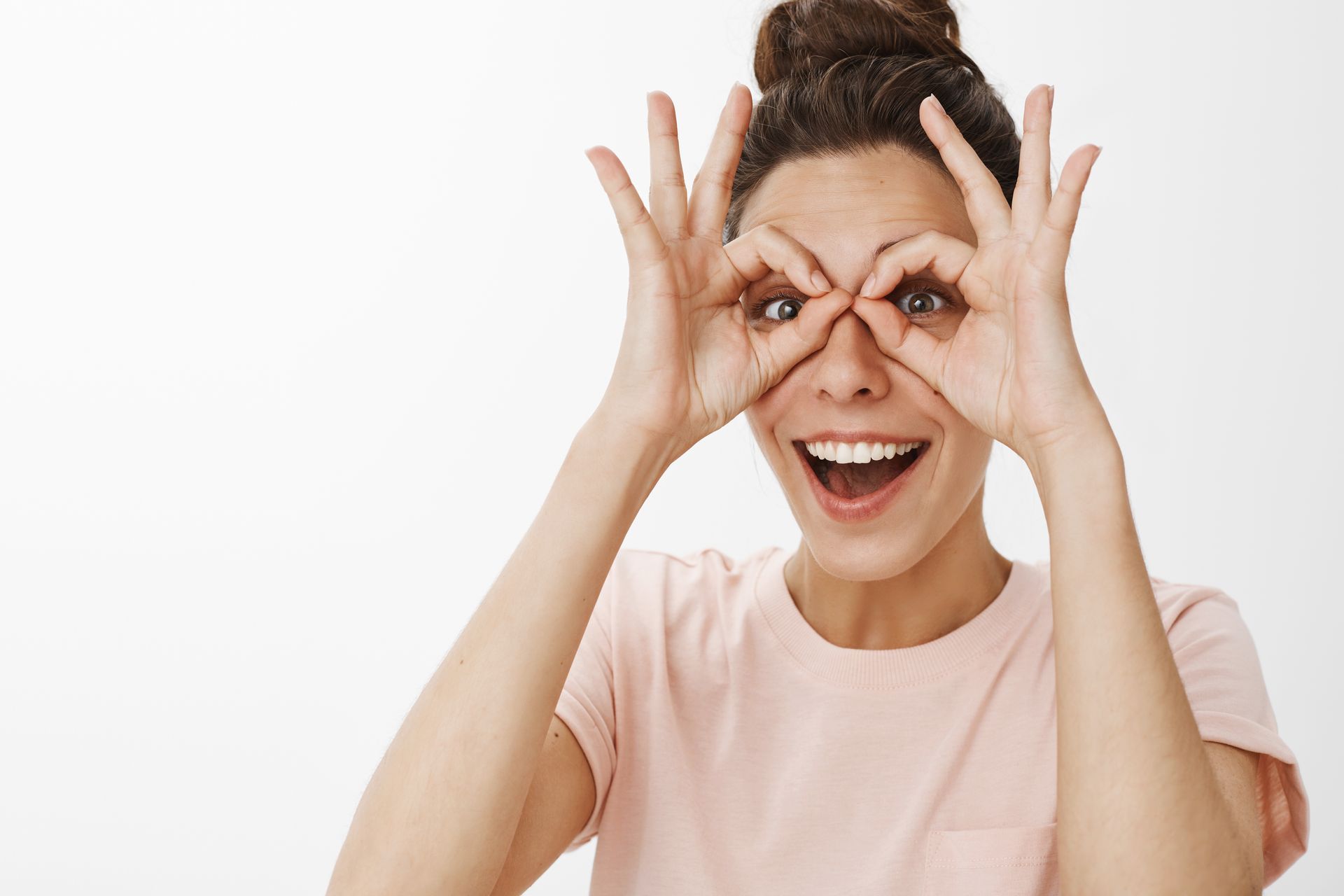 Smiling woman in a light pink shirt making binoculars with her hands against a white background