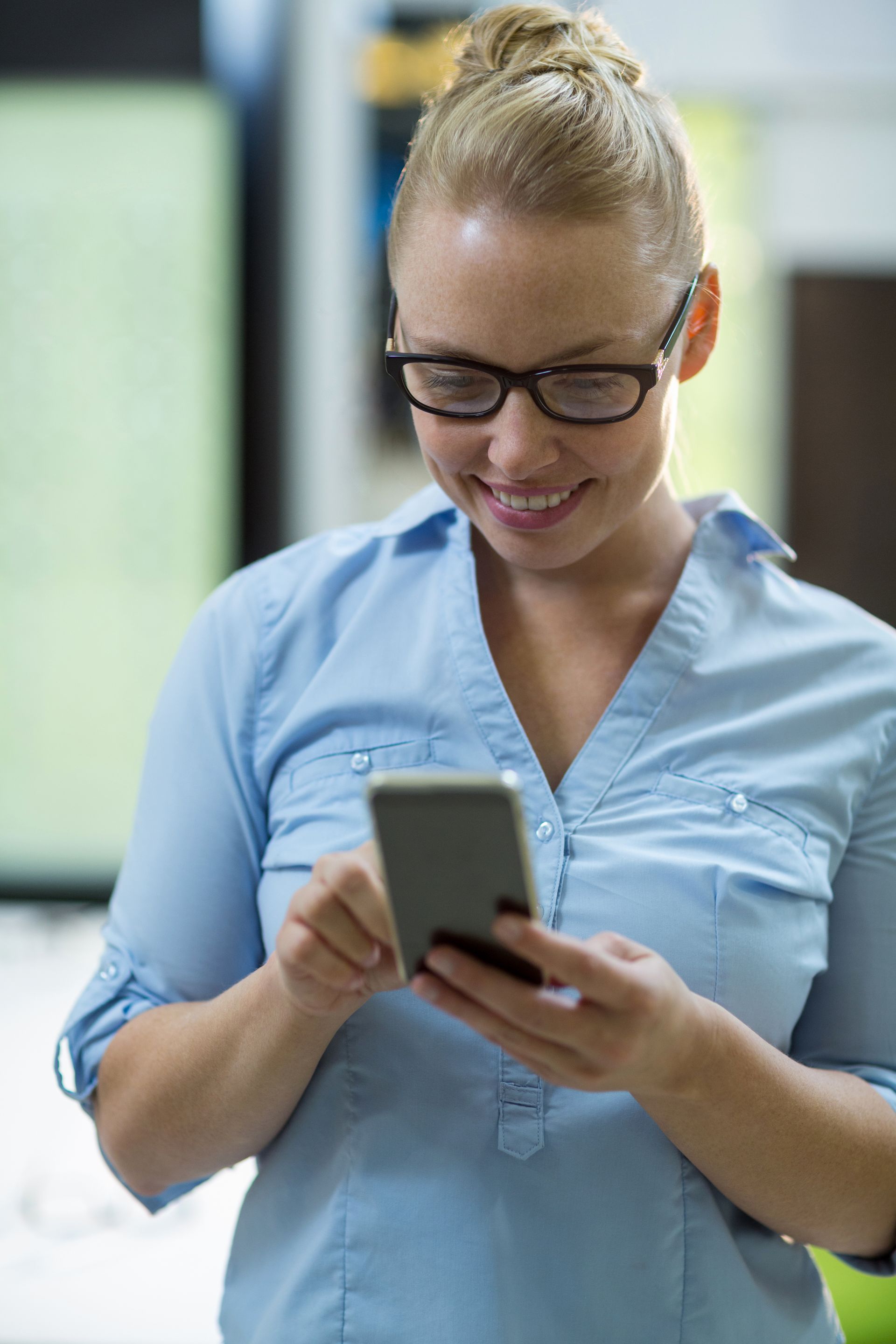 Woman in a light blue shirt smiling while using a smartphone indoors