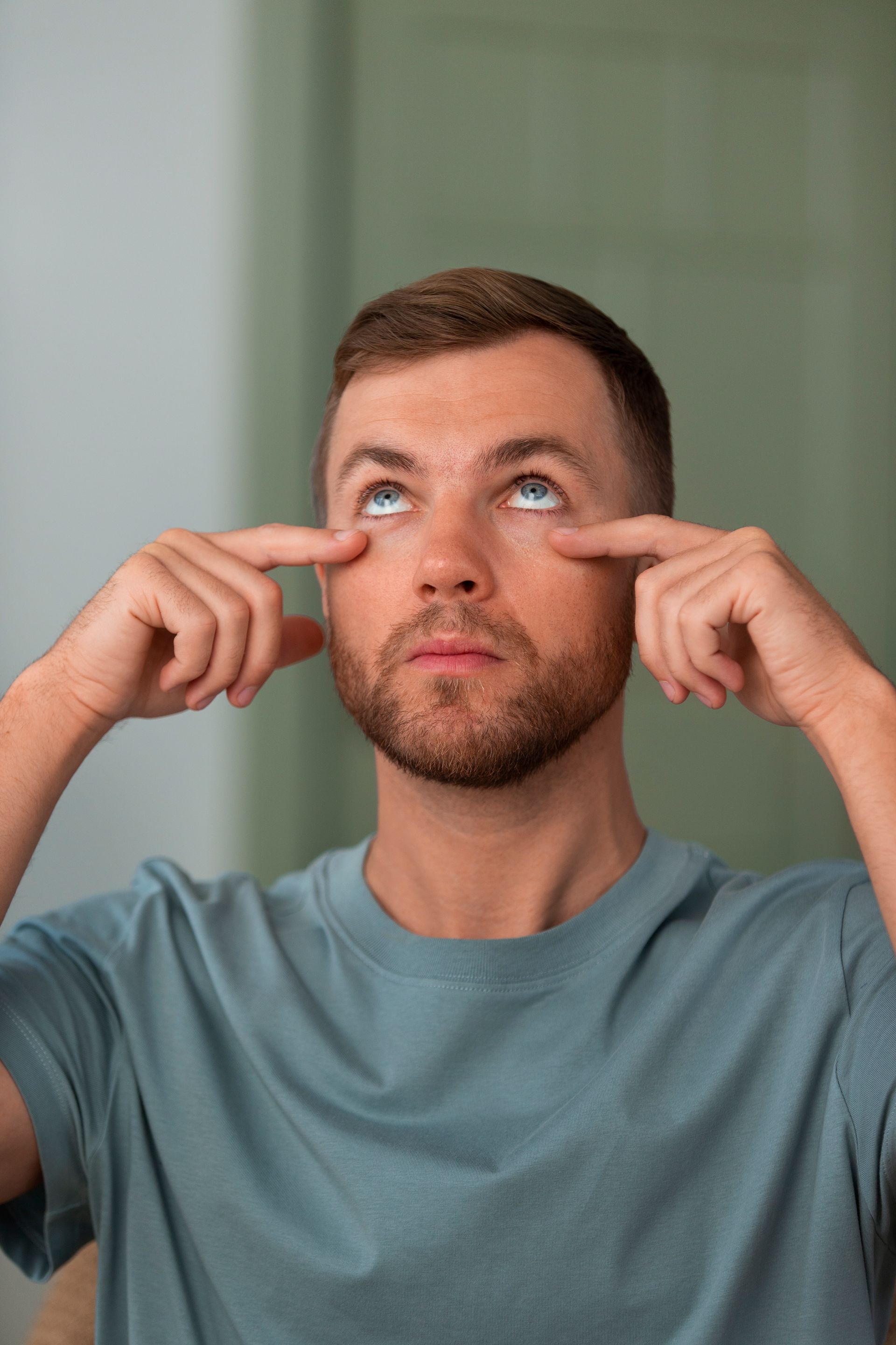 Man in a blue shirt pointing to his eyes while looking upward against a neutral indoor background