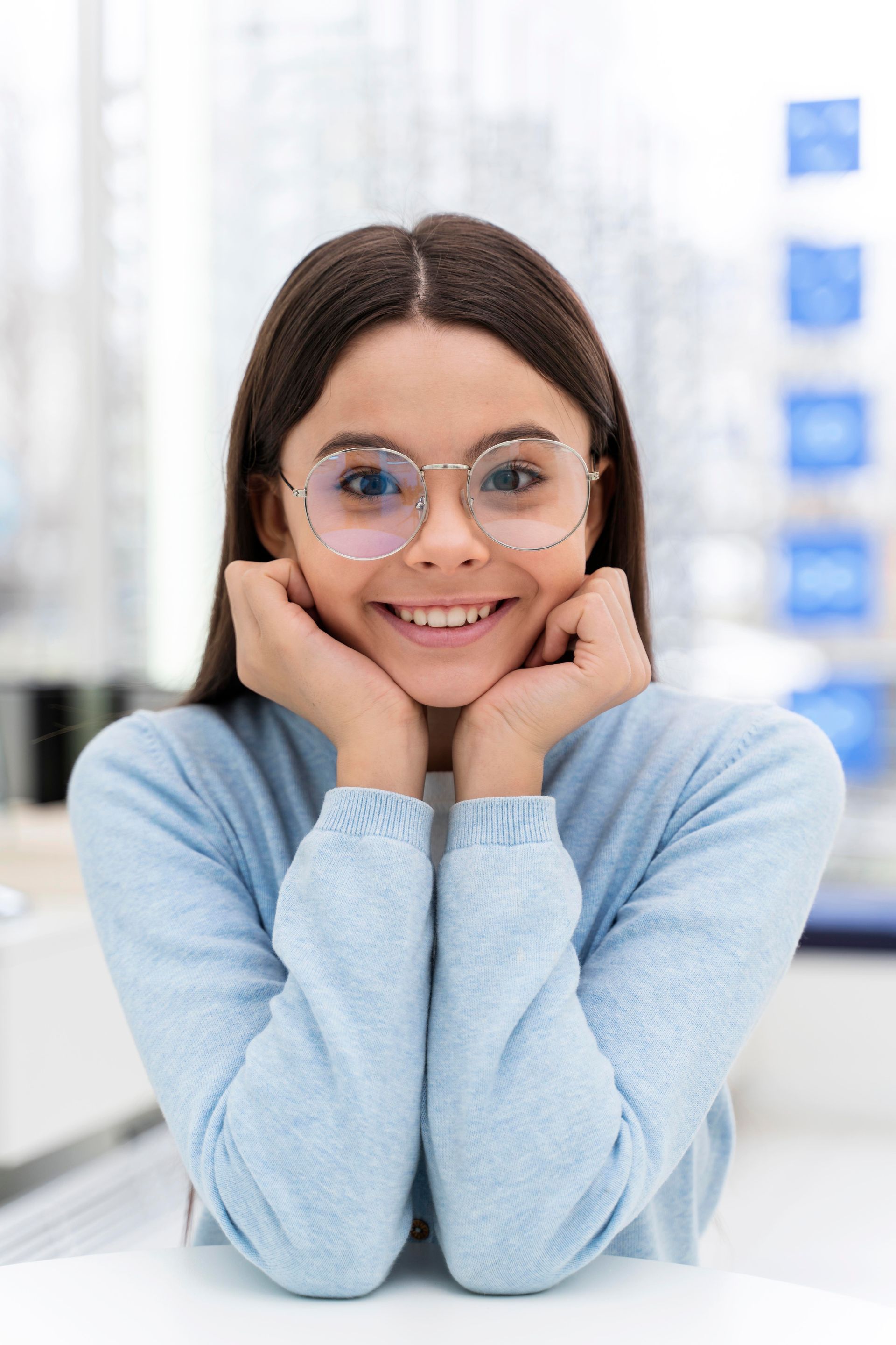 Smiling person with glasses in a light blue sweater, resting chin on hands at a desk indoors