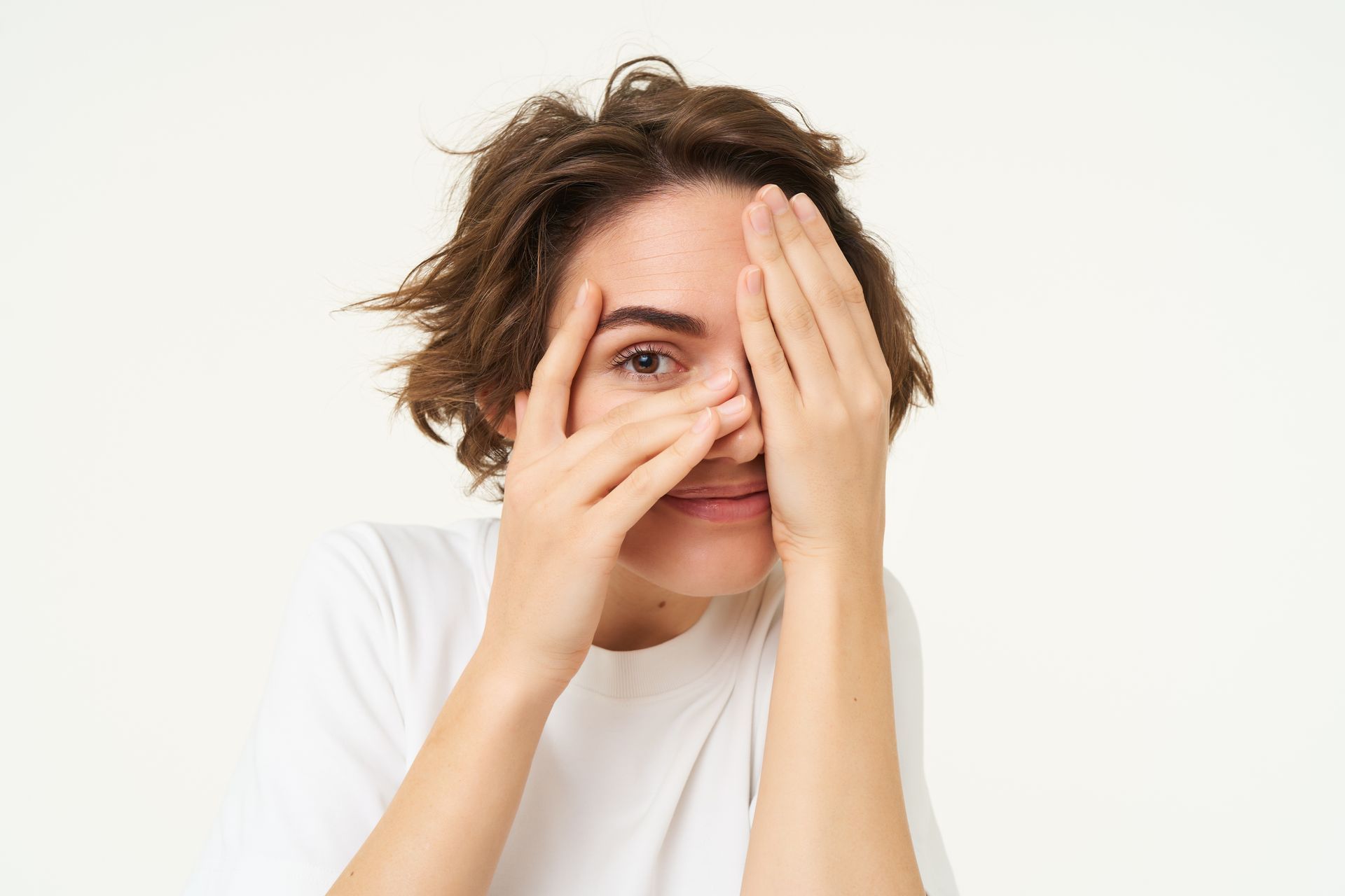 Person peeking through fingers, wearing a white shirt against a plain light background.