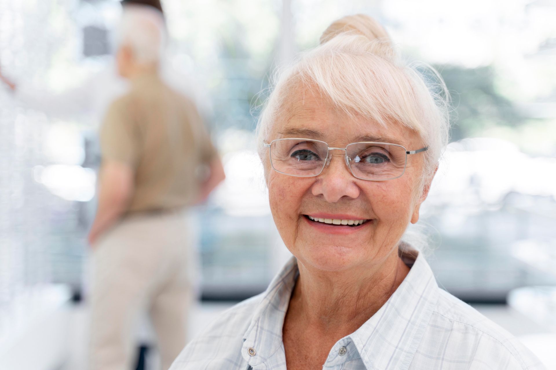 Smiling person with glasses in a bright indoor setting, with a blurred figure in the background