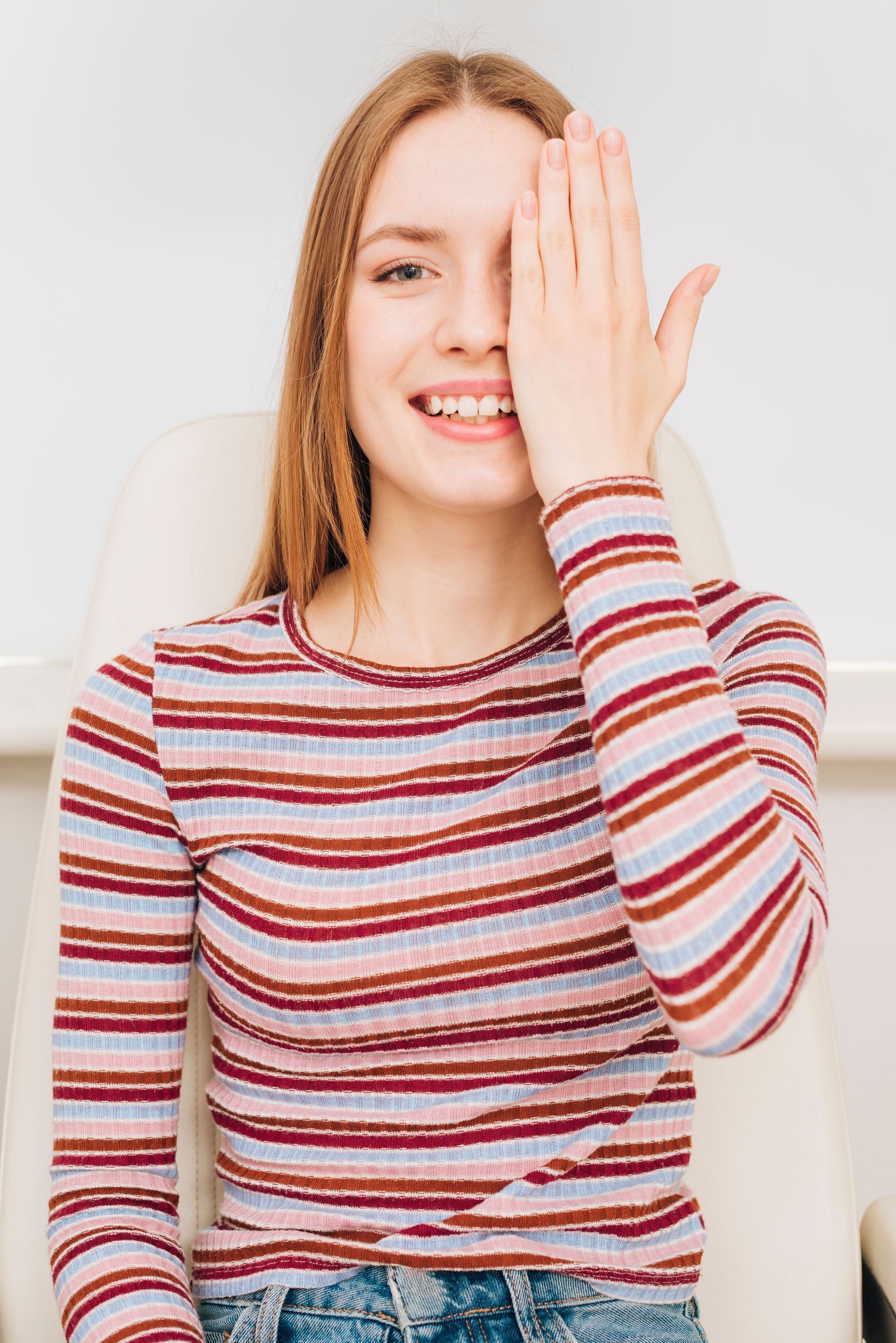Young person in a striped shirt smiling and covering one eye with a hand indoors