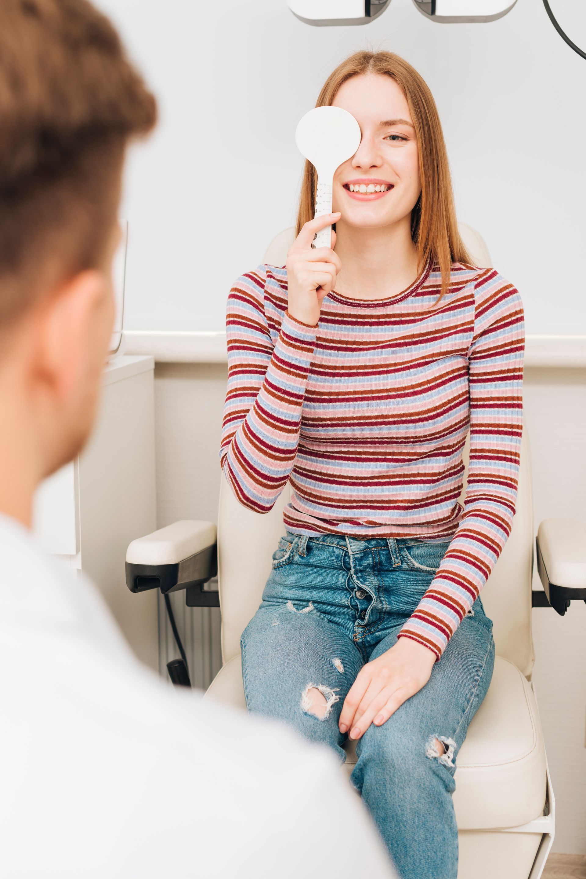 Person holding a small item up to one eye while seated in an exam chair, in a bright clinic room