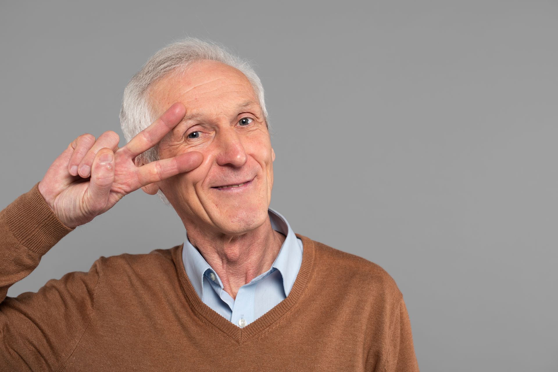 Smiling man in a brown sweater making a peace sign against a gray background