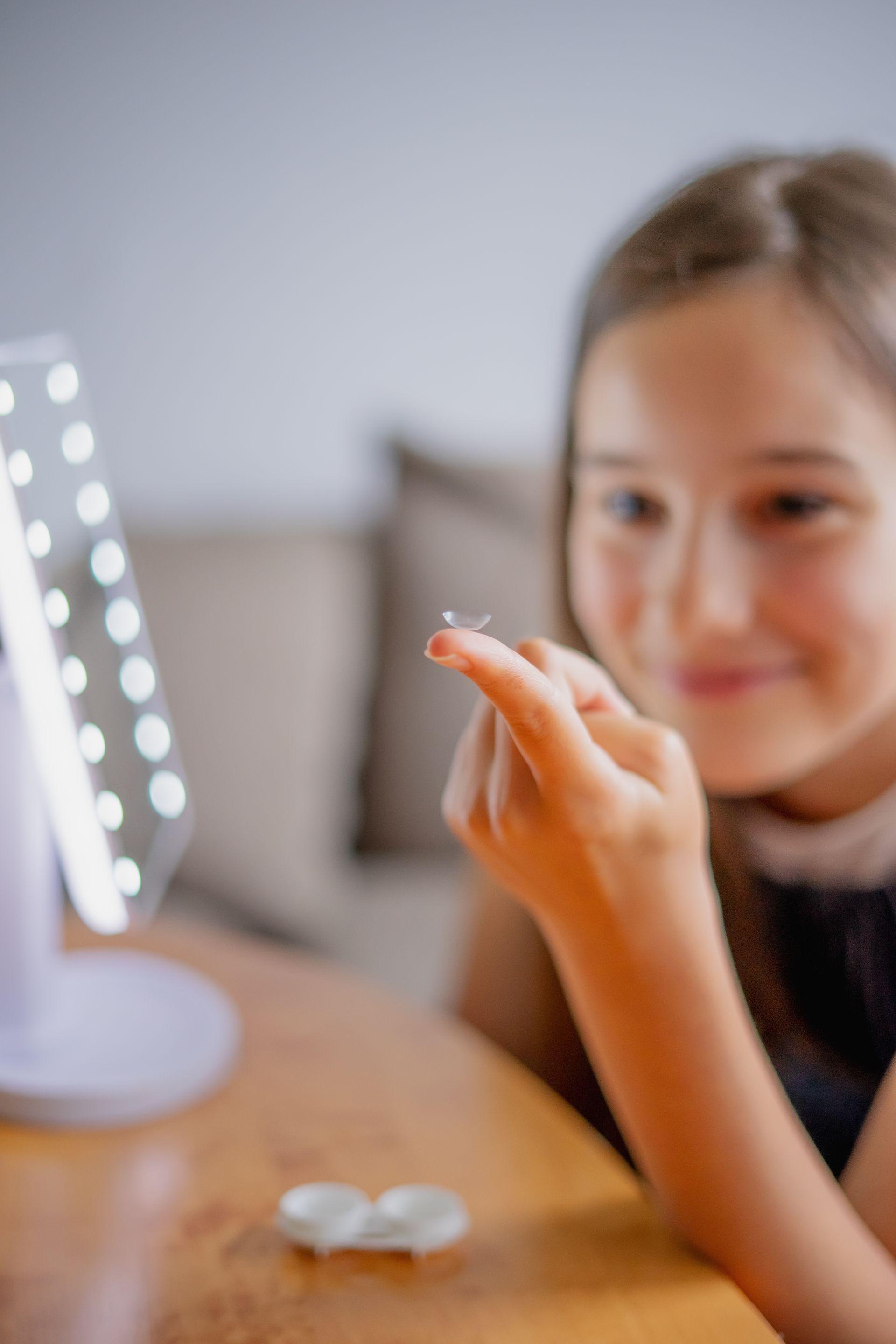 Child applying makeup in front of a lit mirror at a vanity, with a small compact on the table