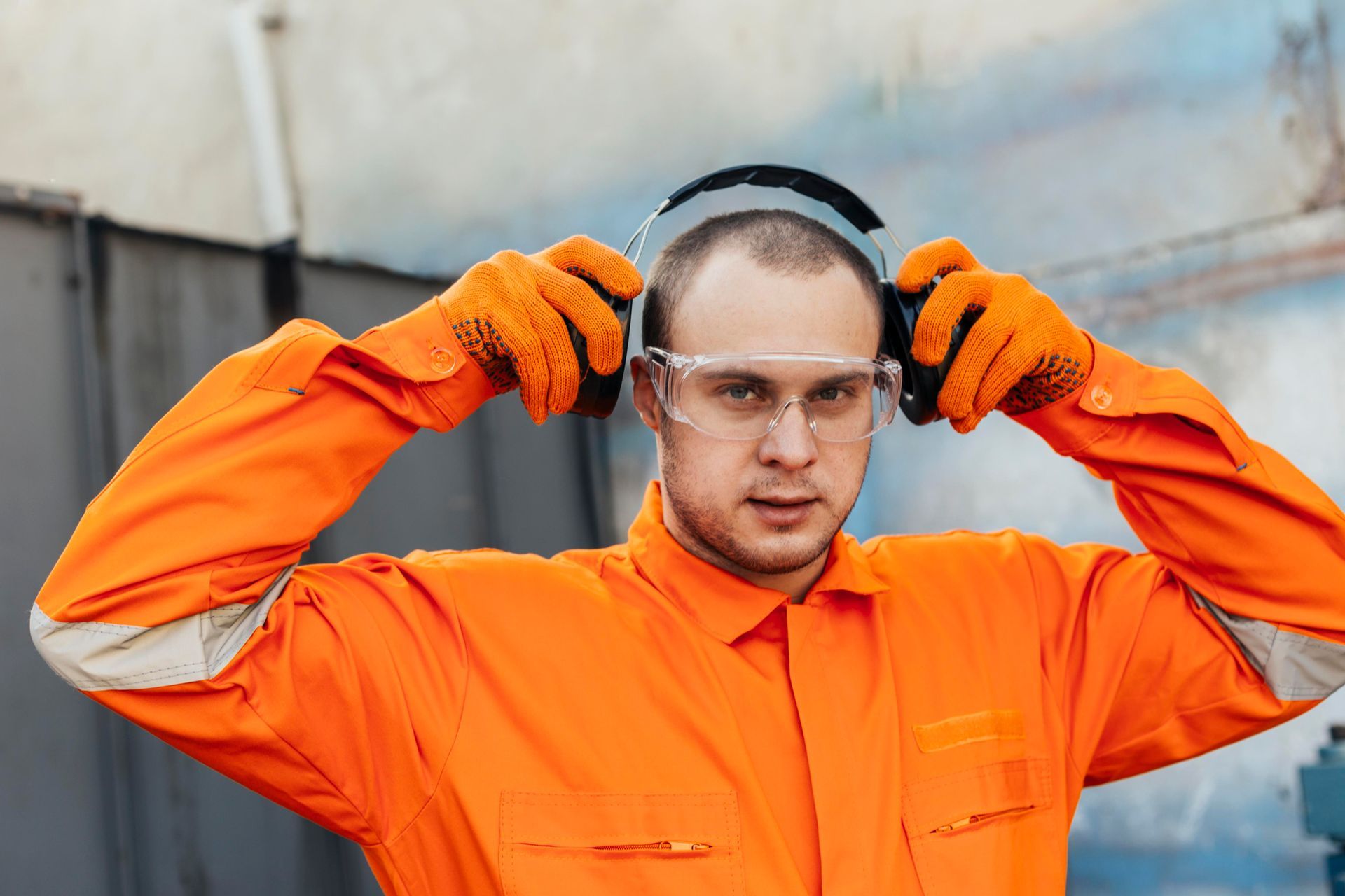Worker in bright orange safety gear adjusting earmuffs and glasses in an industrial setting