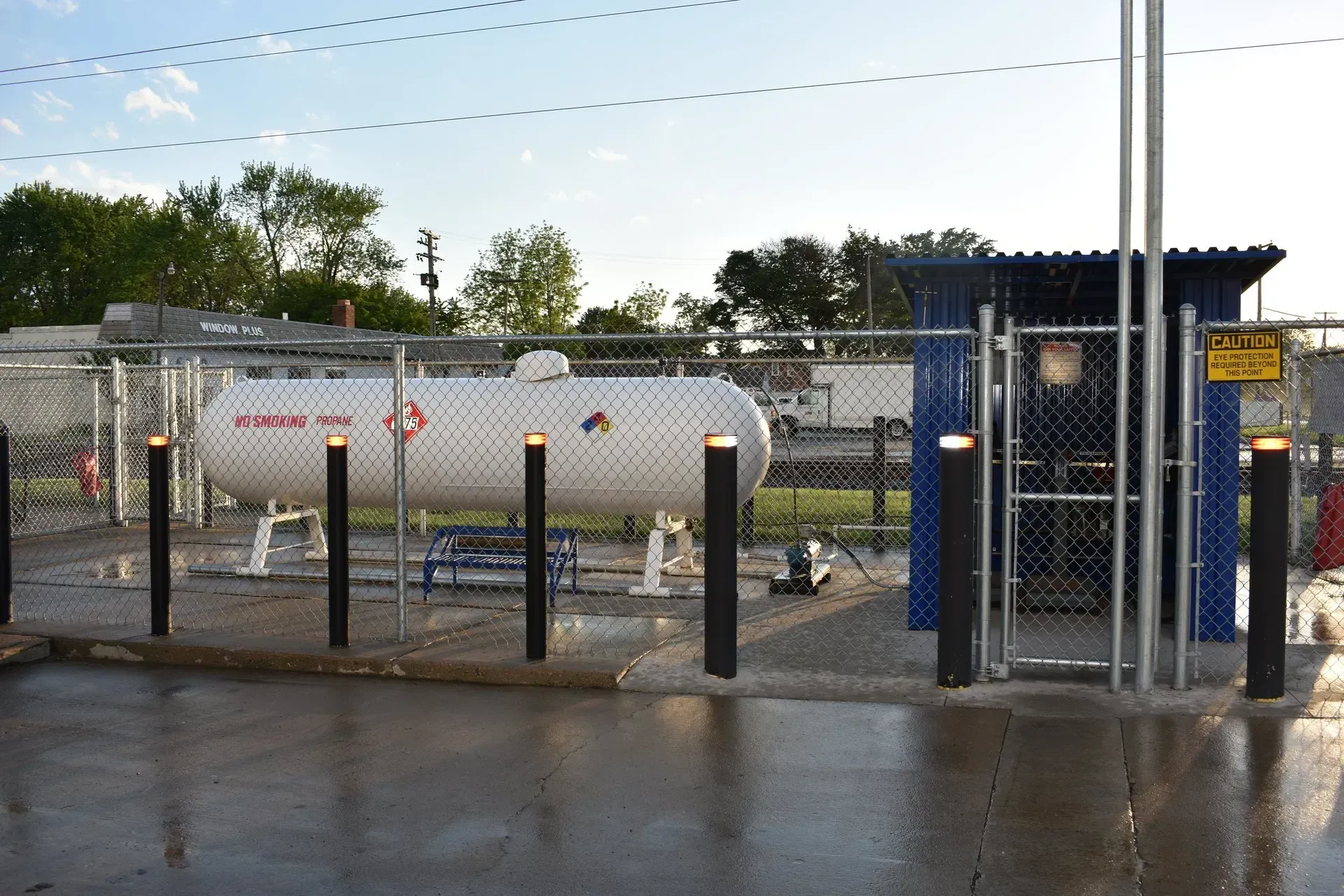 Industrial storage yard with a white propane tank behind a blue gate and black posts on wet pavement.