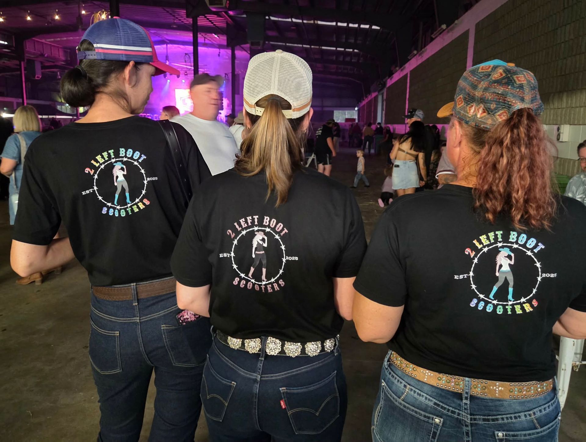 Three people in black shirts stand at a concert, seen from behind, under purple stage lights.
