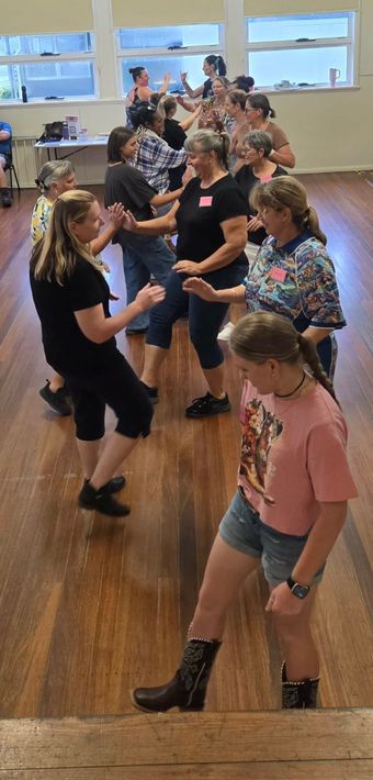 People dancing in a studio with wooden floors and large windows, some wearing black and patterned clothes.