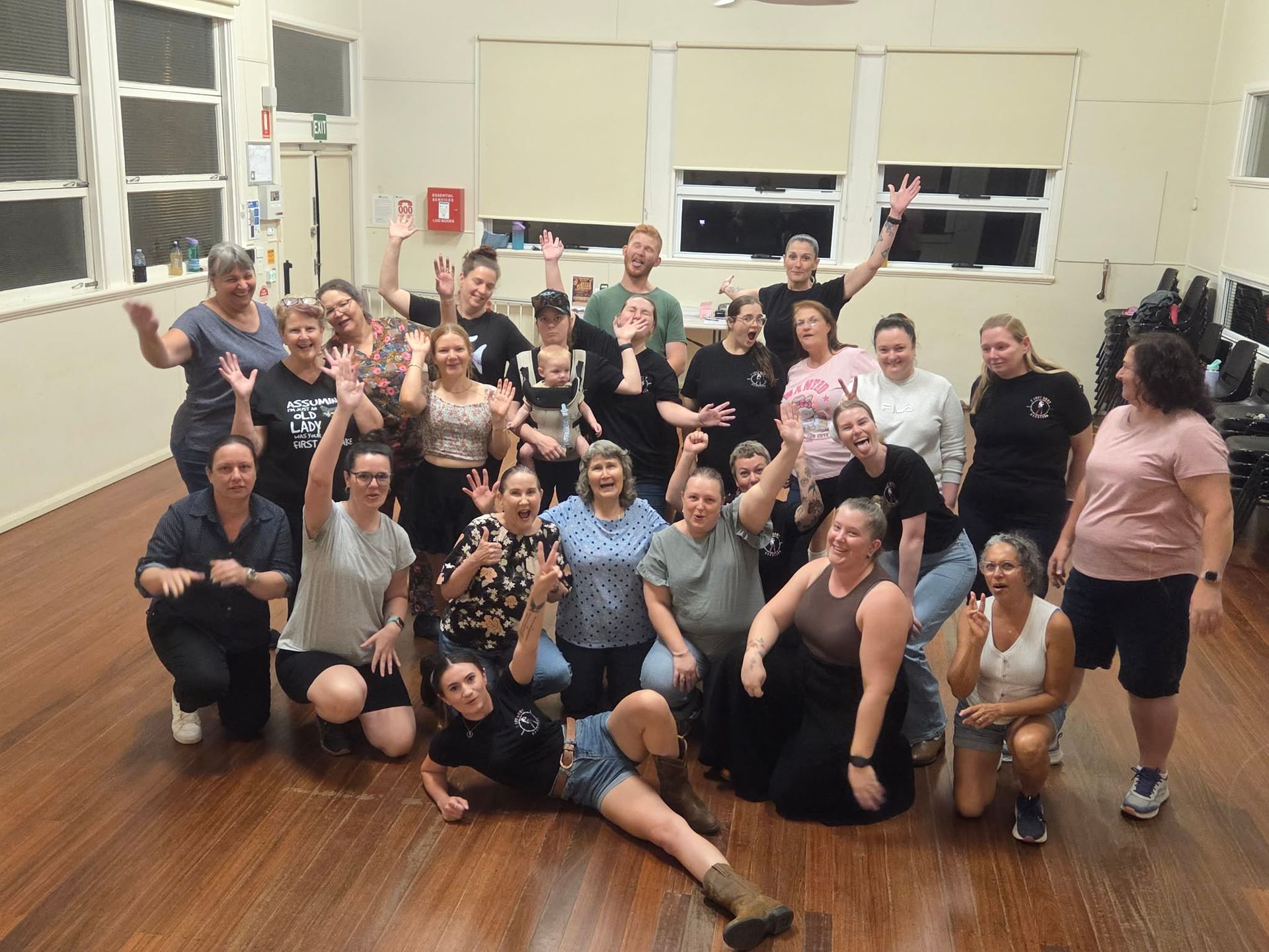 Group of people posing and smiling in a dance studio with wooden floors, some kneeling and waving.