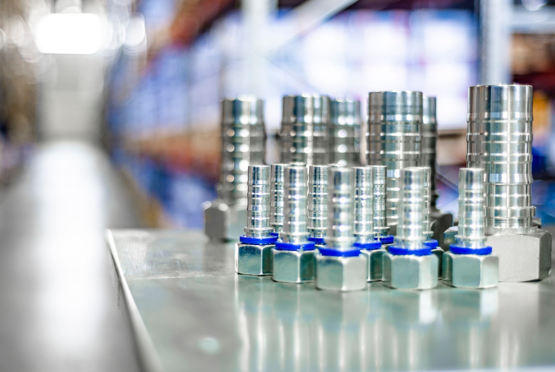 Several metal hydraulic fittings with blue rings arranged on a reflective surface, blurred background of shelves.