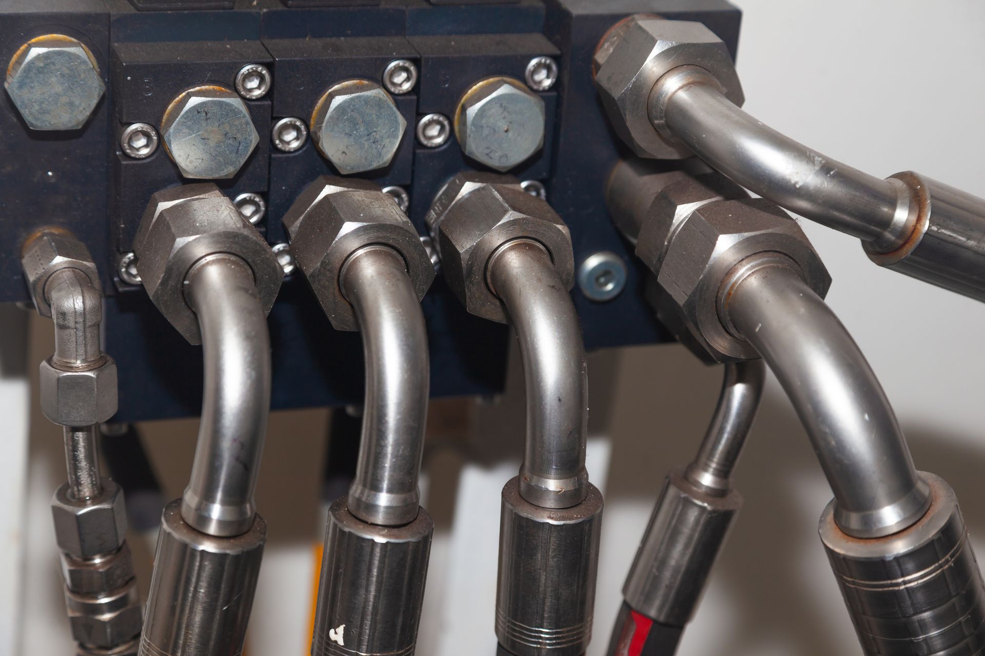 Close-up of hydraulic hoses connected to a black metal block with bolts. Stainless steel tubes curve and connect to fittings.