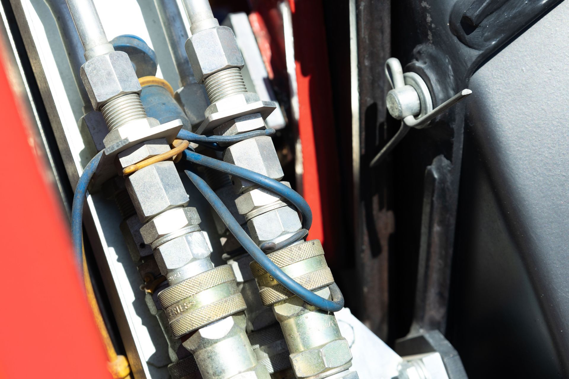Hydraulic fittings with blue wire and rubber band securing lines on red machinery near black trim. Hydraulic fittings with blue wire and rubber band securing lines on red machinery near black trim.