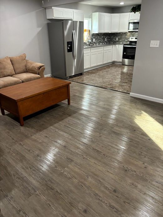 A living room with wood-look flooring, sofa, and a wooden trunk. The kitchen is in the background.