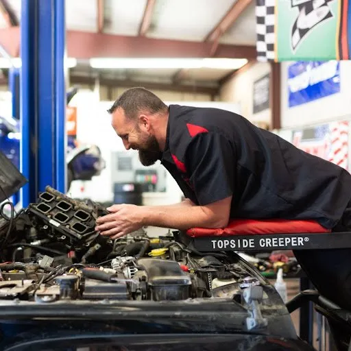 A man is working on a car engine while sitting on a topside creeper