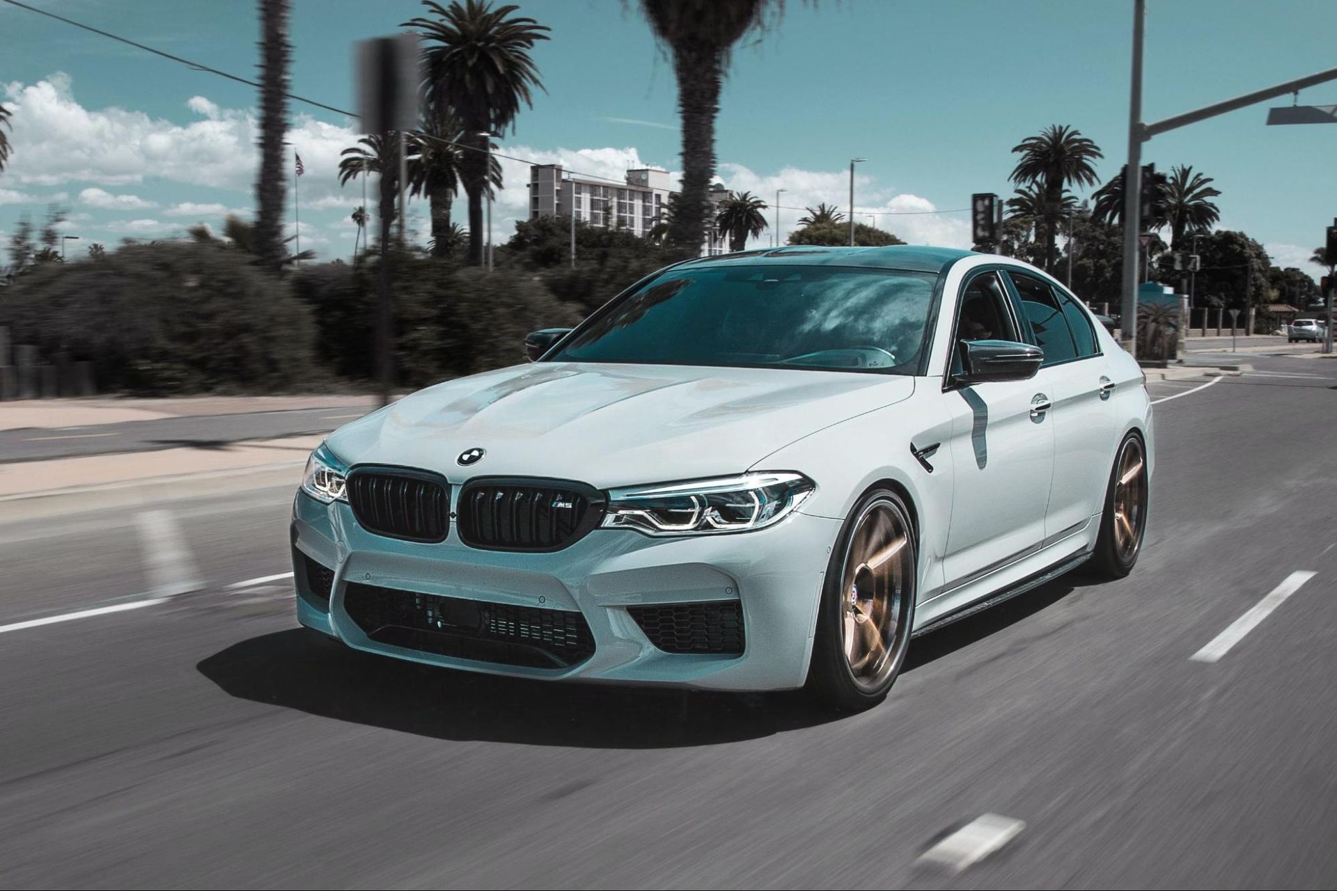 A white bmw m5 is driving down a street with palm trees in the background.