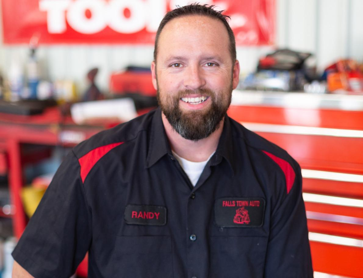 Man in black mechanic uniform with red accents, smiling in auto shop. Name tag reads